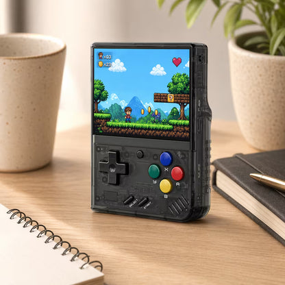 iGameConsole MIYOO Mini Plus transparent black retro portable console on a clean modern desk setup with a notebook, coffee mug, plant, and colorful pixel game screen.