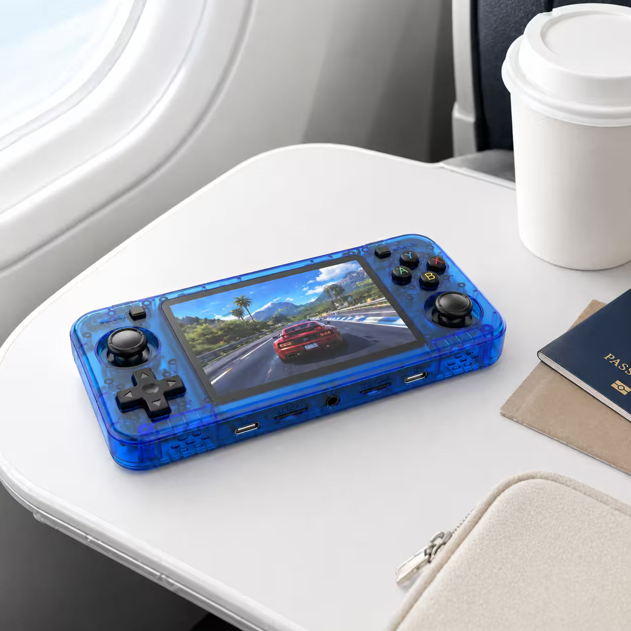 iGameConsole R36H transparent blue portable gaming console on an airplane tray table with a coffee cup, passport, and travel pouch, showing gaming on the go.