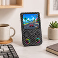 iGameConsole R36T MAX carbon fiber black retro portable console on a clean modern desk setup with a keyboard, coffee mug, notebook, and plant, displaying a colorful racing game screen.