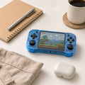 iGameConsole R46H transparent blue compact retro handheld console beside everyday carry items including a notebook, pen, coffee cup, earbuds case, and soft fabric, showing portable size and retro gaming style.