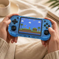 Adult hands holding the iGameConsole R46H transparent blue portable handheld console, showing its compact horizontal design, comfortable grip, dual joysticks, D-pad, ABXY buttons, and retro platform game screen.