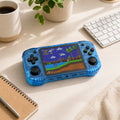 iGameConsole R46H transparent blue retro portable console on a clean modern desk setup with a coffee mug, keyboard, notebook, plant, and colorful pixel game screen.