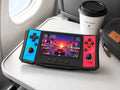 iGameConsole R50 Ultra black portable gaming console with cyan and red side panels on an airplane tray table with a coffee cup and travel pouch, showing gaming on the go.