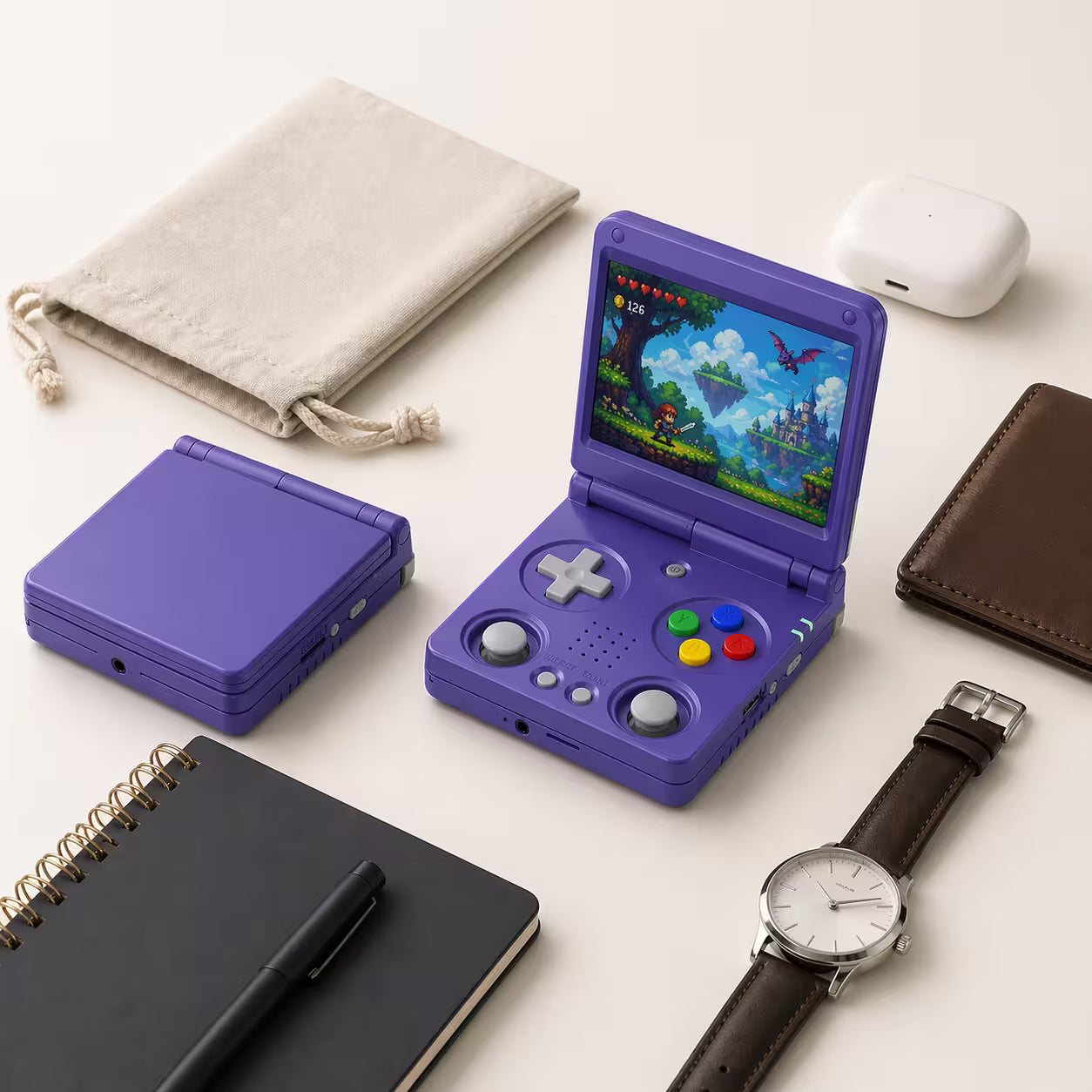 iGameConsole RG34XXSP Foldable Pro purple compact retro handheld console beside everyday carry items including a pouch, notebook, pen, wallet, watch, and earbuds case, showing portable size and foldable design.
