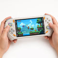 Adult hands holding the iGameConsole RG52 Pro Max off-white and orange portable handheld console, showing its ergonomic grip, horizontal design, dual RGB-ring joysticks, gray D-pad, and action game screen.